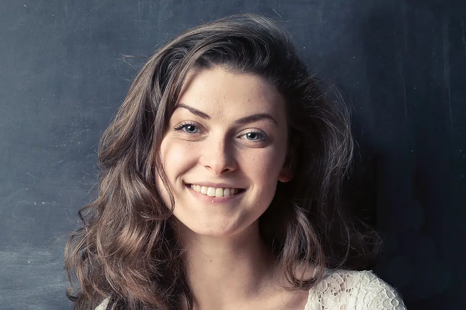 Porträt einer jungen Frau mit einem freundlichen Lächeln. Sie hat braune, wellige Haare und trägt ein weißes Spitzenoberteil. Der Hintergrund ist dunkel, was den Fokus auf ihr Gesicht lenkt und ihren strahlenden Teint hervorhebt. Ihr Blick scheint selbstbewusst und zugänglich, was eine positive und warme Atmosphäre schafft.